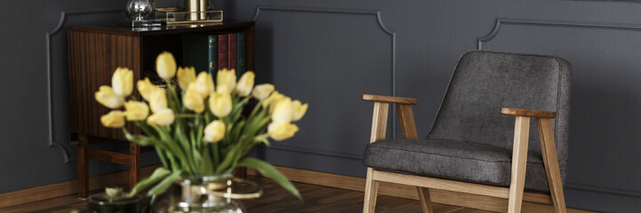 A modern gray armchair in a dark living room interior with wooden furniture and a bouquet of flowers. Real photo.