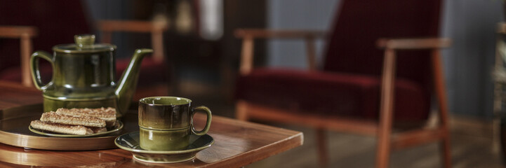 A tea pot, cup and biscuits on a plate on a wooden table and blurry background with dark red armchairs in an elegant living room interior. Real photo.