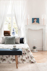 Green bench in front of patterned bed with pillows in white bedroom interior with poster. Real photo