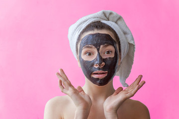 Beauty portrait of a smiling brunette woman in a towel on the head applying black nourishing mask on face on pink background isolated. Skincare cleansing spa relax cosmetics concept
