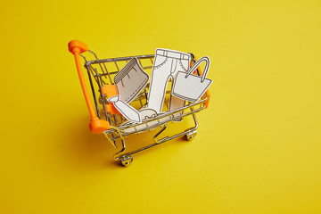 close up view of little shopping cart with clothes made of paper on yellow background