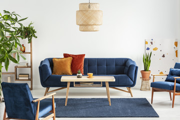 Blue wooden armchairs and couch in living room interior with plants and lamp above table. Real photo