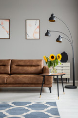 Lamp and table with sunflowers next to leather couch in grey flat interior with posters. Real photo