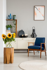 Sunflowers on wooden stool near blue armchair in grey living room interior with poster. Real photo