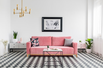 Pillows on pink sofa in spacious living room interior with checkered floor plants and poster. Real photo