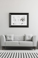 Black and white poster hanging on the wall above grey settee with pillows in real photo of living room interior with striped carpet