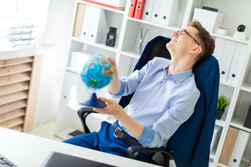 A young man sits in the office at the computer desk, closes his eyes and points a finger at the globe.
