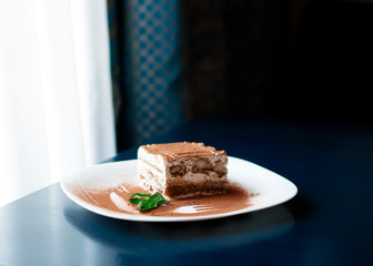 Tiramisu traditional Italian dessert on white plate on dark blue table next to roon window with curtains . Withe day light from the window. Close up food with fork and knife decor