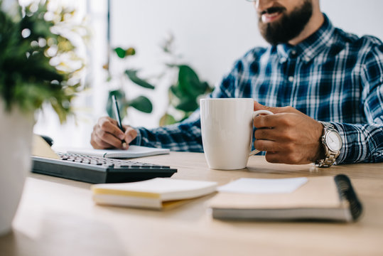 cropped shot of successful smiling seo with cup of coffee and computer writing notes at workplace