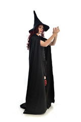 full length portrait of red haired girl wearing long black cloak, pointy hat and witch costume. standing pose, isolated on white studio background.