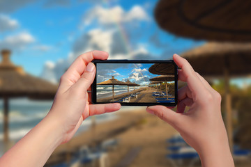 Female taking picture on mobile phone of double rainbow over ocean and tropical beach with umbrellas chairs and tables.