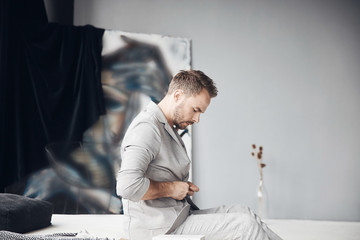 Handsome man with dark hair and beard fastens his light jacket and sits on the bed in scandinavian room