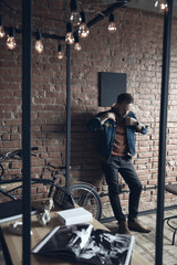 Man in denim jacket stands near brick wall in loft styled interior with bicycle and yellow lamps