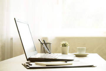 Blank screen laptop computer, cup of cappuccino coffee, cactus, supplies and folded eye glasses on wooden desk in spacious office full of sunlight. Creative workspace. Close up, copy space, background