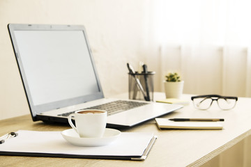 Fototapeta premium Blank screen laptop computer, cup of cappuccino coffee, cactus, supplies and folded eye glasses on wooden desk in spacious office full of sunlight. Creative workspace. Close up, copy space, background