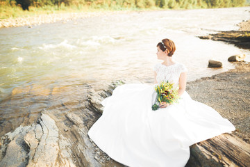 Gorgeous beautiful bride smiles while posing by the river with bouquet