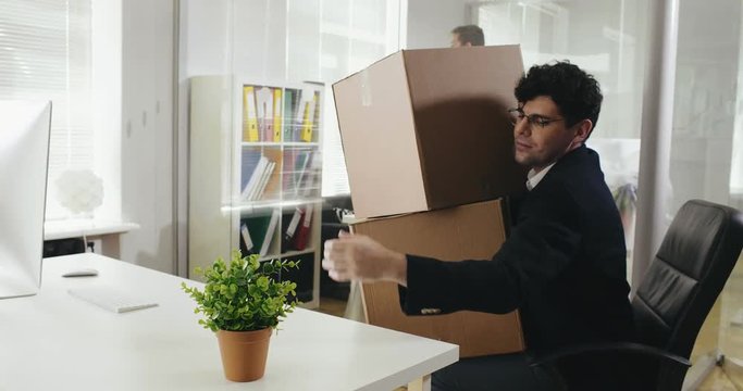 Young specialist got fired and leaving his office. European man carrying boxes with stuff and plant and leaving his workplace after dismissal.
