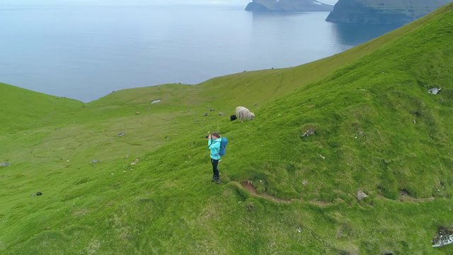 AERIAL: Woman photographer hiking near the Kallur lighthouse taking photos of the breathtaking ocean off a grassy mountain. Flying around young female tourist photographing the scenic countryside.