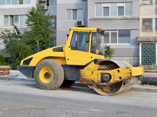 Yellow combination asphalt compactor at the site of road construction site near the residential building, side view, summer day