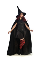 full length portrait of red haired girl wearing long black cloak, pointy hat and witch costume. standing pose, isolated on white studio background.