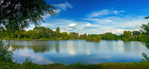 Fototapeta premium Tranquil landscape at a lake, with white clouds sky and the trees reflected in the clean blue water