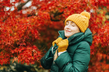 Autumn portrait of young beautiful woman wearing green jacket and set of yellow hat and gloves, posing next to bright red Japanese maple tree. Eyes closed