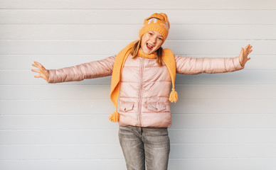 Expressive child posing against white background, wearing soft pink padded jacket and set of orange knitted hat and scarf. Winter fashion for children - obrazy, fototapety, plakaty