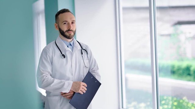 Handsome confident doctor in white coat looking at window and holding clipboard in his hands