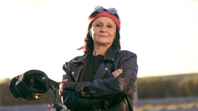 An Elderly Woman In A Leather Jacket And Gloves Stands In The Background Of An Empty Road. The Woman Smiles And Puts Her Hands On Each Other, Turns Sideways . Happy Life Of An Elderly Rocker And Biker