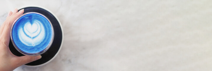Banner of top view of a woman's left hand grabbing a cup of hot butterfly pea latte with beautiful heart-shaped latte art on a light marble table background, morning refreshment with copy space 