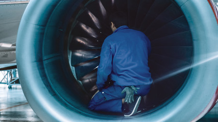 Aircraft maintenance mechanic with a flash light inspects plane turbine blades in a hangar.