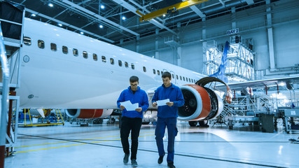 Two aircraft maintenance mechanics have a conversation while checking documents in a plane hangar with an airplane in the background.
