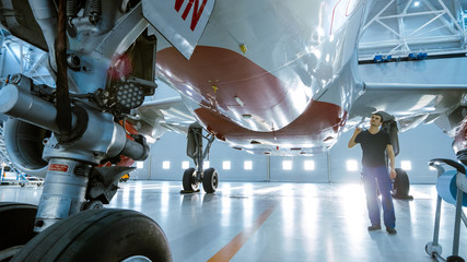 In a Hangar Aircraft Maintenance Engineer/ Technician/ Mechanic Visually Inspects Airplane's Chassis and Body/Fuselage walking Under It. - obrazy, fototapety, plakaty