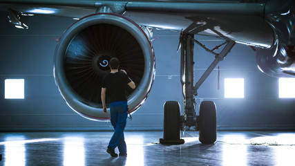 In a Hangar Aircraft Maintenance Engineer/ Technician/ Mechanic Inspects with a Flashlight Airplane's Jet Engine.