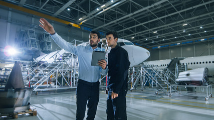 Aircraft Maintenance Worker and Engineer having Conversation. Holding Tablet and Points with His...