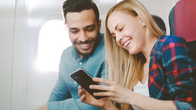 On a Board of Commercial Airplane Beautiful Young Blonde with Handsome Hispanic Male Watch Social Media on Smartphone and Laugh. Sun Shines Through Aeroplane Window.
