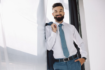 smiling businessman holding jacket and standing near window in office