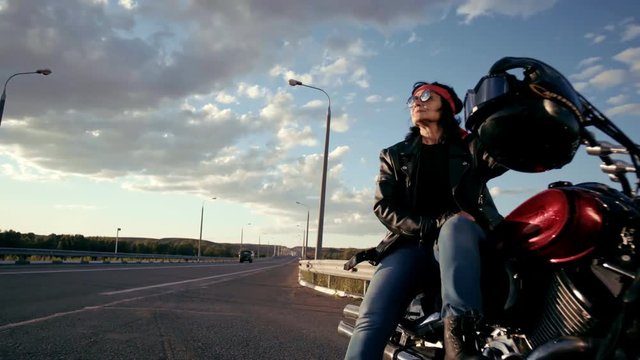 A Biker Old Woman In A Leather Jacket And Gloves Is Sitting On Her Cool Motorcycle. A Woman In Round Glasses And A Red Bandana. There's An Empty Highway In The Background. The Life Of An Elderly