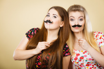 Obraz premium Two happy women holding fake moustache on stick