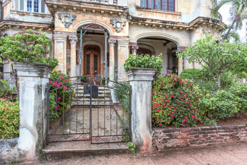fence with bushes and a metal gate to a dilapidated old mansion with columns
