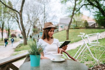 Attractive woman with hat sitting in outdoor cafe and using tablet.