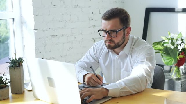 concentrated attractive muscular man in glasses and white shirt working on notebook and using graphic tablet in modern office drawing 3D design illustration studio young successful art manager inside