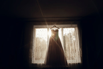 beautiful wedding dress hanging on hanger on window in morning light.