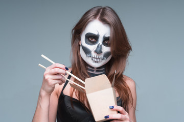 Young beautiful girl with a sugar skull make up, holds in her hand a paper box for instant noodles and chopsticks. Halloween face art on gray background.