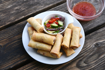Fried Spring Rolls Thai style in white dish with cucumber salad and sweet chili sauce on wooden floor