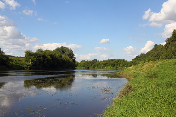 Beautiful picturesque calm small river with green forest shore in summer against the blue sky with clouds and reflection in the water