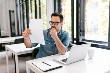 Serious young businessman reading paperwork at desk in office.