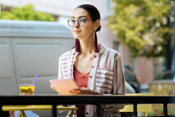 Good day. Beautiful calm woman sitting at the cafe terrace and smiling while holding a modern tablet