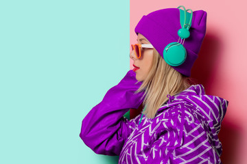 Young blonde girl in 90s sports jacket and hat on pink and blue background.