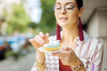 Holding an eclair. Calm relaxed woman sitting in a cafe and looking at the tasty eclair on the plate